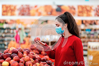 A young woman in a medical mask on her face looking at apples in a supermarket. The concept of shopping and the new Stock Photo