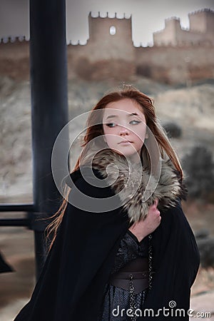 A young woman in a medieval dress walks through a field in a warm winter cloak with fur. medieval palace in the background Stock Photo