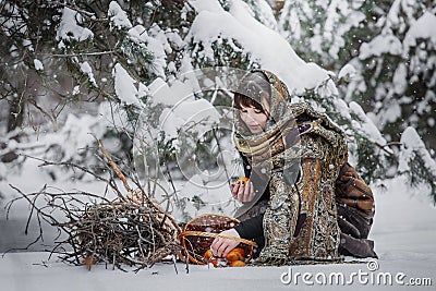 A young woman in old clothes is sitting with brushwood and a basket with apples in the snow in the winter forest Stock Photo