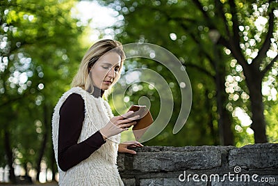 Young woman portrait in summer. Blonde girl is reading message on cell phone outside in city nature. Female with telephone. Stock Photo