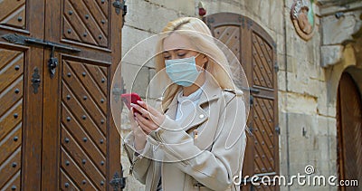 Young woman in protective mask using smartphone in the street of old city. Worried girl searching news about Covid-19 Stock Photo