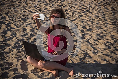 Young woman in red dress with computer and smartphone on the beach. Freelance and downshifting concept. Stock Photo