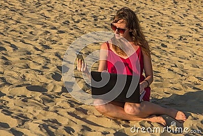 Young woman in red dress with computer and smartphone on the beach. Freelance and downshifting concept. Stock Photo