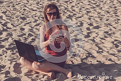 Young woman in red dress with computer and smartphone on the beach. Freelance and downshifting concept. Stock Photo