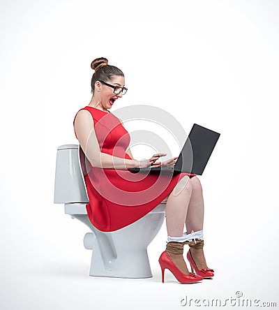 Young woman in red dress and glasses working on laptop sitting on toilet Stock Photo