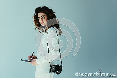 Young woman with red hair holding notebook and pen while standing with digital camera on grey Stock Photo