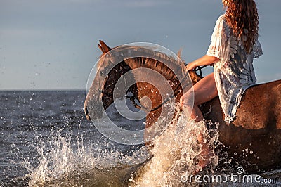 a woman riding a brown horse into the ocean waves of the shore Stock Photo