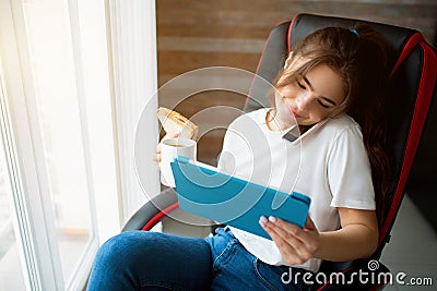 Young woman in room during quarantine. Businesswoman in chair talking on phone and look at tablet with smile. Hold Stock Photo