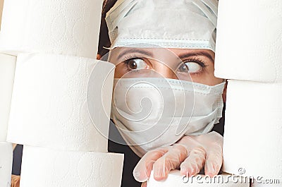 Young woman with scared eyes in two medical virus protection face masks looks through stacks of toilet paper. Covid-19 quarantine Stock Photo