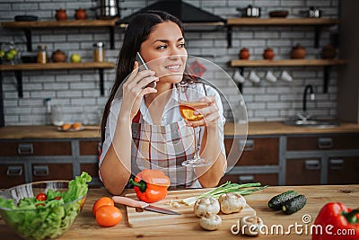 Young woman sit at table in kitchen. She drink white wine nd talk on phone. Woman looks happy. Vegetables lying on desk. Stock Photo