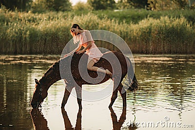Woman sits astride a horse that drinks water on background of water splashes Stock Photo