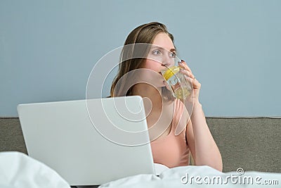 Young woman sitting in bed with laptop computer and drinking water with lemon Stock Photo