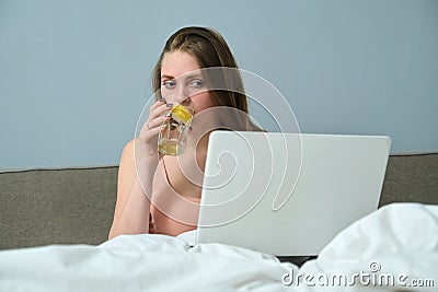 Young woman sitting in bed with laptop computer and drinking water with lemon Stock Photo