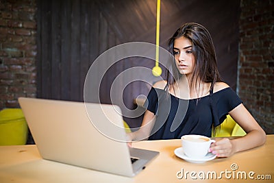 Young woman sitting in coffee shop at wooden table, drinking coffee and using smartphone. On table is laptop. Girl browsing intern Stock Photo