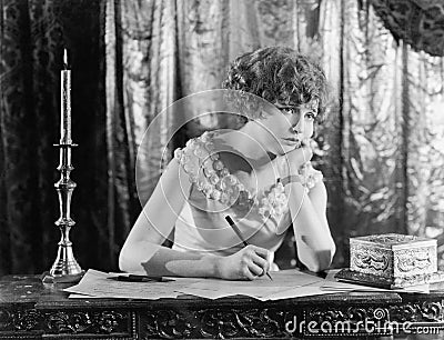 Young woman sitting at a desk with a pen in hand, looking sad while writing a letter Stock Photo