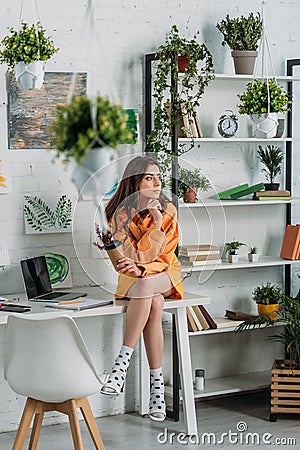 Young woman sitting on desk in room decorated with green plants and paintings on wall Stock Photo