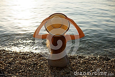 Young woman is sitting on the pebble shore with beach dress on holding a straw hat on her head. Back view portrait of Stock Photo