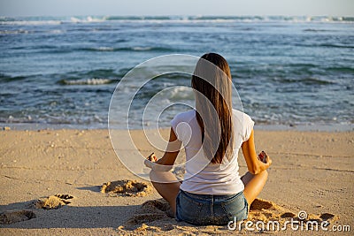 Young woman sitting on the sand in lotus pose in front of the ocean. Yoga at the beach. Hands in gyan mudra. Meditation concept. Stock Photo