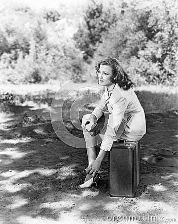 Young woman sitting on the side of the road on her suitcase, rubbing her aching feet Stock Photo