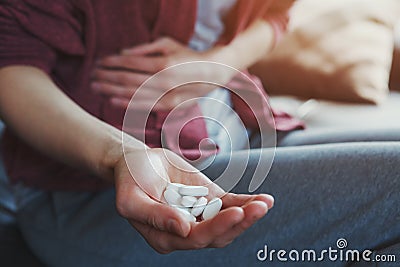 Young woman sitting on sofa with hand on aching stomach holding white medicine pills Stock Photo