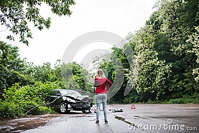 A young woman with smartphone by the damaged car after a car accident, making a phone call. Stock Photo