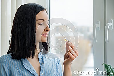 Young woman standing at home near the window taking vitamin Omega 3 fish oil yellow capsule Stock Photo
