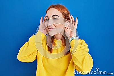 Young woman standing over blue background trying to hear both hands on ear gesture, curious for gossip Stock Photo
