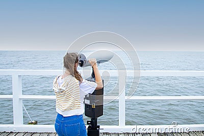 A young woman stands on the pier, looking through coin binoculars. Stock Photo