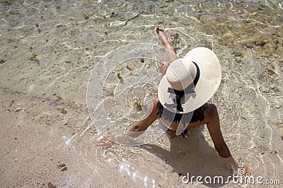 Young woman in a stylish hat and a two-piece bikini sitting in shallow turquoise water Stock Photo