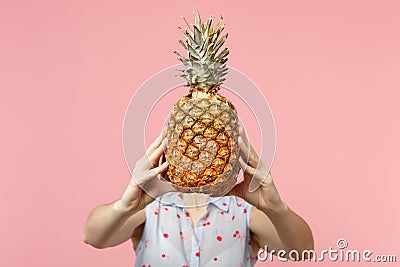 Young woman in summer clothes hold in hands hiding covering face with fresh ripe pineapple fruit on pink pastel Stock Photo