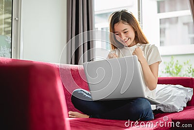 Young woman surfing the internet by laptop on red sofa and glad in successful trading in her house. Selling and online shopping Stock Photo