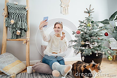 Young Woman in sweater taking selfie photo while relaxing with cat on floor cusions near potted christmas tree in modern Stock Photo