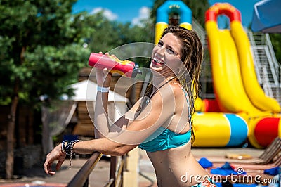 Young woman in a swimsuit drinks water from a bottle. Charming tanned athletic woman stands in the sun in a swimsuit and drinks Stock Photo