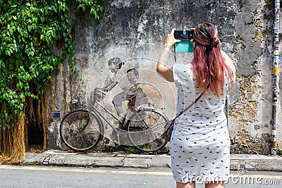 Young woman taking a photo of Street Art mural boy and girl on bicycle on a wall in George Town on Penang island in Malaysia Editorial Stock Photo