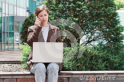 Young woman talking on the phone and typing on a laptop on street. Stock Photo