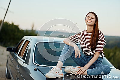 A young woman travels alone in her car on the roads in the countryside and relaxes sitting on the hood watching the Stock Photo