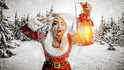 Young woman in a uniform of Santa Claus with a lit lantern on a blurred snowy winter Christmas day background. Stock Photo