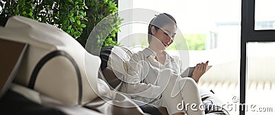 A young woman is using a computer tablet while sitting on a big cushioned frameless chair Stock Photo