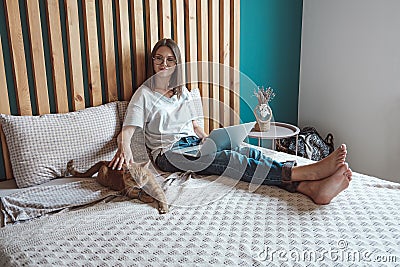 Young woman using laptop to work, lying on the bed with a pet cat in the bedroom, . Flexible hours and remote work. Stock Photo