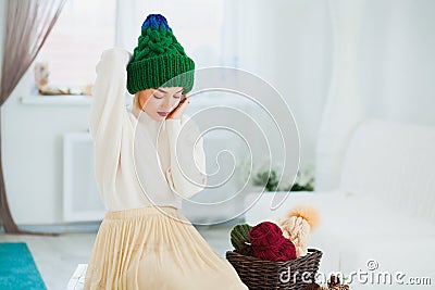 Young woman in warm hand knitted hat at home. Pretty lady in big green cap, smiling happily. Beautiful girl knitting in cozy flat Stock Photo