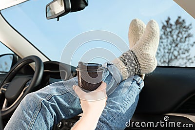 Young woman in warm socks holding her legs on car dashboard and drinking coffee. Stock Photo