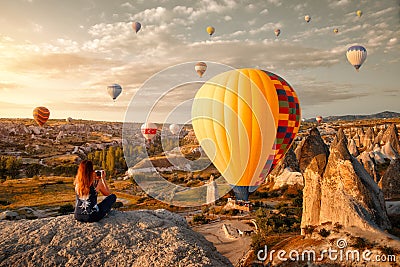 Young woman watches and photographs flying colorful balloons on an early morning in Goreme Valley, Cappadocia. Turkey Stock Photo