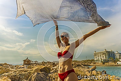 Young woman waving scarf in wind at beach. Happy woman in red bikini holding tissue and looking away at sea. Stock Photo