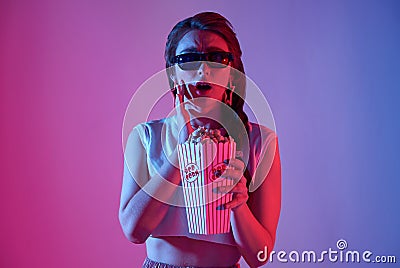 Young woman wearing 3D glasses holding popcorn and beverage watching exciting movie on neon background Stock Photo