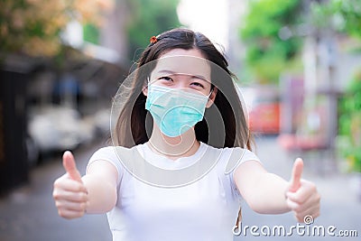 Young woman wearing green medical face mask. Person showing thumbs up and sweet smile under protection mask. Stock Photo