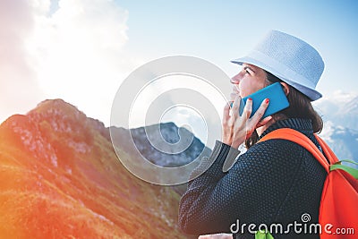 Young woman wearing a hat and backpack talking on the phone, sitting high in mountains at sunset Stock Photo