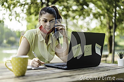 Young woman wearing open protective face mask using laptop outdoors - Female entrepreneur taking notes while calling with cell Stock Photo