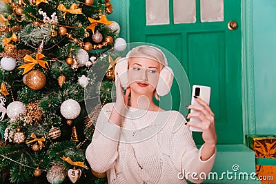 Young woman in white, fluffy headphones in Christmas decorations with a phone in her hands. a smartphone Stock Photo