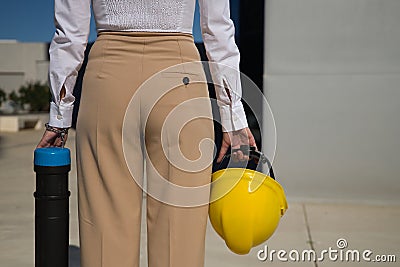 Young woman in white shirt, brown pants, with yellow workman`s helmet and blueprint pipe in hands, back turned, arriving at offic Stock Photo