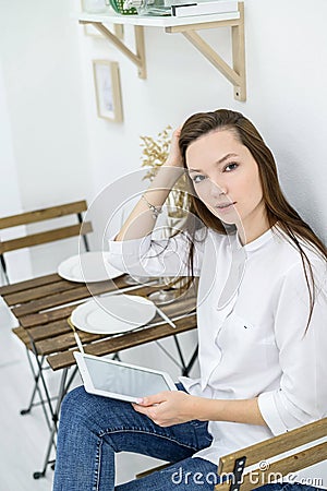 A young woman in a white shirt and jeans sits in a cafe at a table with a tablet in her hands. Female office worker at Stock Photo